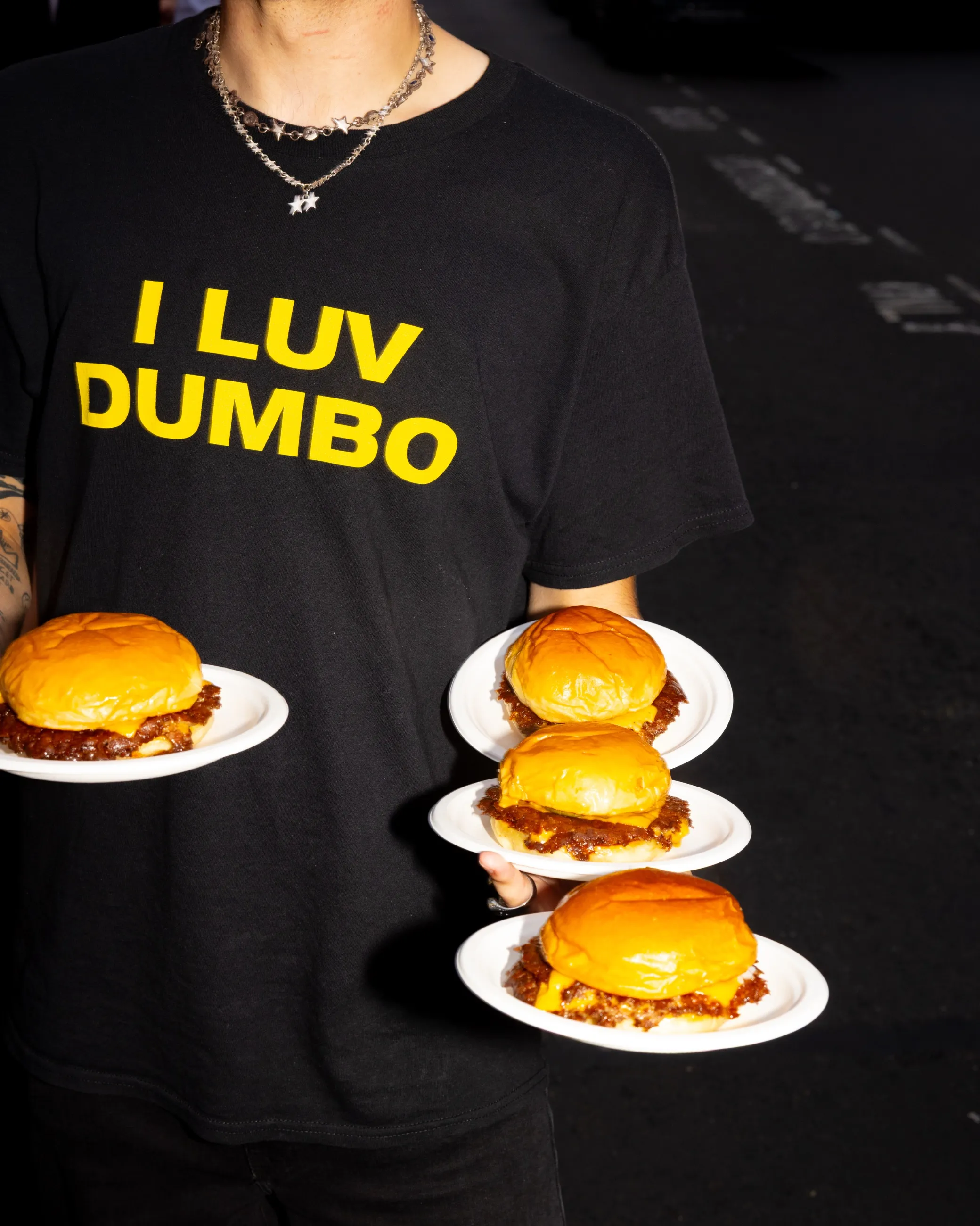 A Dumbo server in an I LUV DUMBO t-shirt carrying four plated cheeseburgers